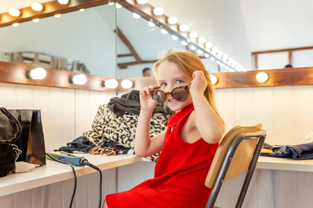 A girl, a child, a model in a beautiful dress and sunglasses sitting near mirror with lamps and smiling. Fashon kid beauty salon photosession conceptの写真素材