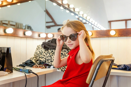 A girl, a child, a model in a beautiful dress and sunglasses sitting near mirror with lamps and smiling. Fashon kid beauty salon photosession conceptの写真素材