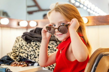 A girl, a child, a model in a beautiful dress and sunglasses sitting near mirror with lamps and smiling. Fashon kid beauty salon photosession conceptの写真素材