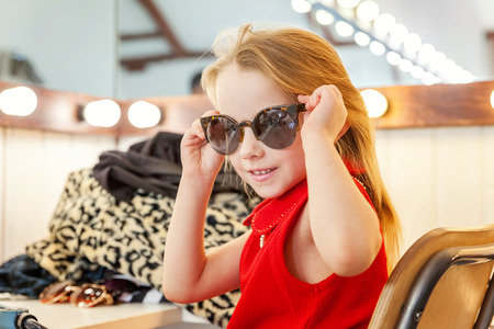 A girl, a child, a model in a beautiful dress and sunglasses sitting near mirror with lamps and smiling. Fashon kid beauty salon photosession conceptの写真素材