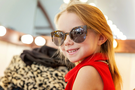 A girl, a child, a model in a beautiful dress and sunglasses sitting near mirror with lamps and smiling. Fashon kid beauty salon photosession conceptの写真素材