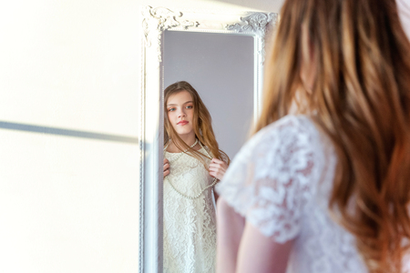 beauty, hygiene, morning and people concept - smiling happy teenage girl looking at reflection in mirror. young positive woman wearing white dress posing in bright light room against white wallの写真素材