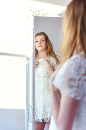 beauty, hygiene, morning and people concept - smiling happy teenage girl looking at reflection in mirror. young positive woman wearing white dress posing in bright light room against white wallの写真素材