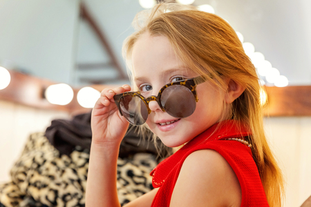 A girl, a child, a model in a beautiful dress and sunglasses sitting near mirror with lamps and smiling. Fashon kid beauty salon photosession conceptの写真素材