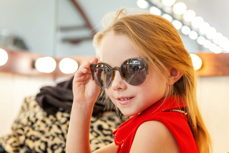 A girl, a child, a model in a beautiful dress and sunglasses sitting near mirror with lamps and smiling. Fashon kid beauty salon photosession conceptの写真素材