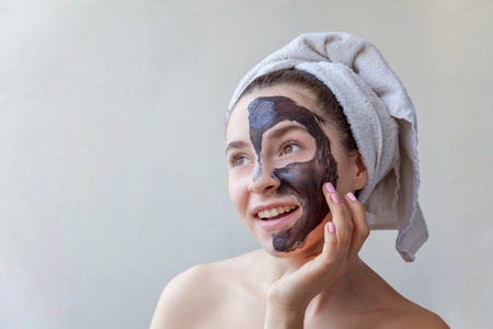 Beauty portrait of a smiling brunette woman in a towel on the head applying black nourishing mask on face on white background isolated. Skincare cleansing spa relax cosmetics conceptの写真素材