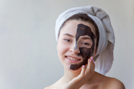 Beauty portrait of a smiling brunette woman in a towel on the head applying black nourishing mask on face on white background isolated. Skincare cleansing spa relax cosmetics conceptの写真素材