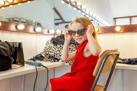 A girl, a child, a model in a beautiful dress and sunglasses sitting near mirror with lamps and smiling. Fashon kid beauty salon photosession conceptの写真素材