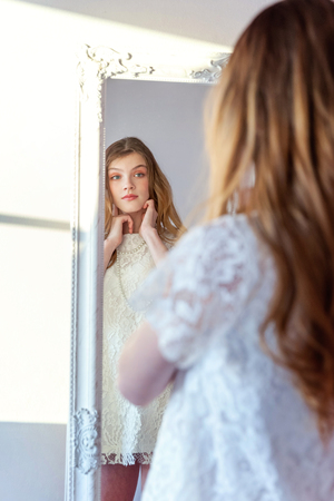 beauty, hygiene, morning and people concept - smiling happy teenage girl looking at reflection in mirror. young positive woman wearing white dress posing in bright light room against white wallの写真素材