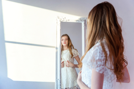 beauty, hygiene, morning and people concept - smiling happy teenage girl looking at reflection in mirror. young positive woman wearing white dress posing in bright light room against white wallの写真素材