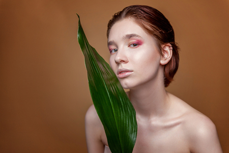 Young beauty brunette woman with fashion make up and green leaves on natural brown background. Youth teens cleansing eco organic cosmetic relax and skin care concept. Spa and wellnessの写真素材