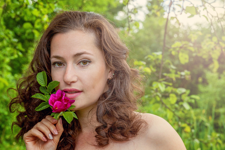 Beautiful young brunette girl with perfect skin holding red rose flower in hand on park or garden green background. Beauty nature skincare eco organic cosmetic ecology pure health conceptの写真素材
