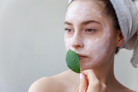 Beauty portrait of a smiling brunette woman in a towel on the head with white nourishing mask or creme on face and green leaf in hand on white background isolated. Skincare cleansing eco organic cosmetic spa relax conceptの写真素材