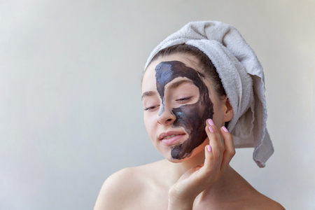 Beauty portrait of a smiling brunette woman in a towel on the head applying black nourishing mask on face on white background isolated. Skincare cleansing spa relax cosmetics conceptの写真素材