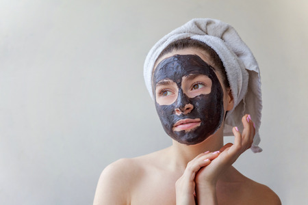 Beauty portrait of a smiling brunette woman in a towel on the head applying black nourishing mask on face on white background isolated. Skincare cleansing spa relax cosmetics conceptの写真素材