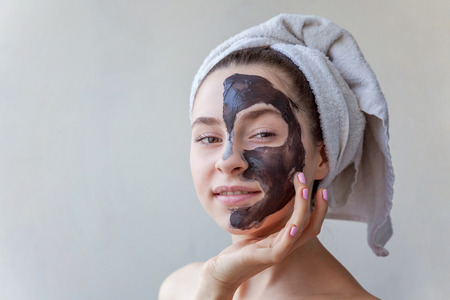 Beauty portrait of a smiling brunette woman in a towel on the head applying black nourishing mask on face on white background isolated. Skincare cleansing spa relax cosmetics conceptの写真素材
