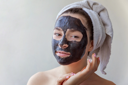 Beauty portrait of a smiling brunette woman in a towel on the head applying black nourishing mask on face on white background isolated. Skincare cleansing spa relax cosmetics conceptの写真素材