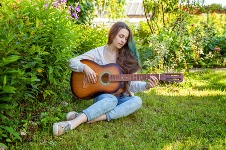 Young hipster woman sitting in grass and playing guitar on park or garden background. Teen girl learning to play song and writing music. Hobby, lifestyle, relax, Instrument, leisure, education conceptの写真素材