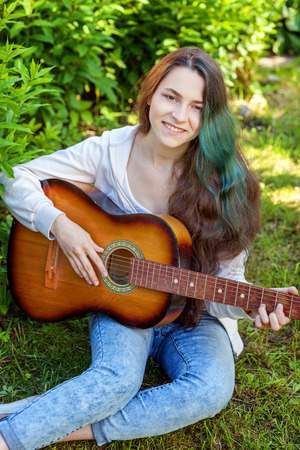 Young hipster woman sitting in grass and playing guitar on park or garden background. Teen girl learning to play song and writing music. Hobby, lifestyle, relax, Instrument, leisure, education conceptの写真素材