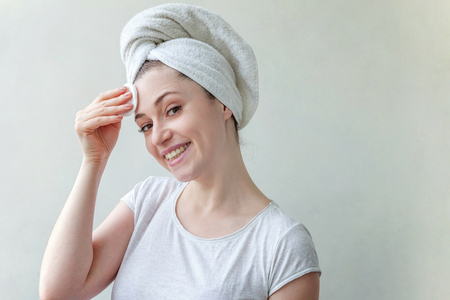 Beauty portrait of a smiling brunette woman with towel on head with soft healthy skin removing make up with cotton pad isolated over white background. Skincare cleansing spa relax conceptの写真素材