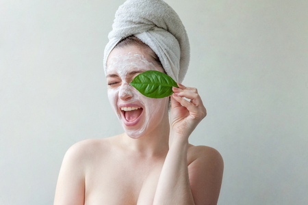 Beauty portrait of a smiling brunette woman in a towel on the head with white nourishing mask or creme on face and green leaf in hand on white background isolated. Skincare cleansing eco organic cosmetic spa relax conceptの写真素材