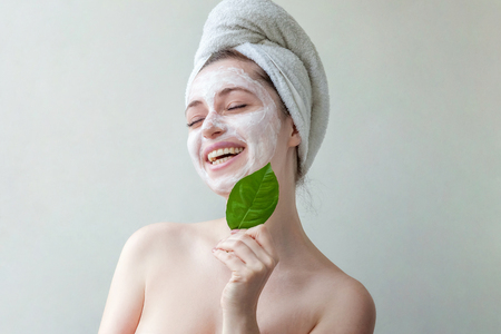 Beauty portrait of a smiling brunette woman in a towel on the head with white nourishing mask or creme on face and green leaf in hand on white background isolated. Skincare cleansing eco organic cosmetic spa relax conceptの写真素材