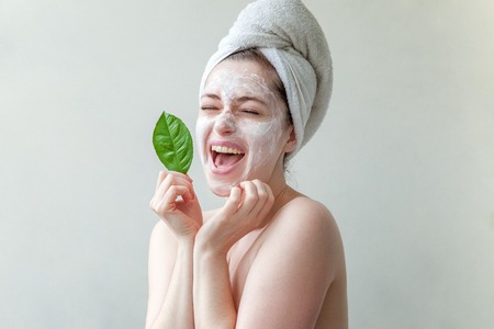 Beauty portrait of a smiling brunette woman in a towel on the head with white nourishing mask or creme on face and green leaf in hand on white background isolated. Skincare cleansing eco organic cosmetic spa relax conceptの写真素材