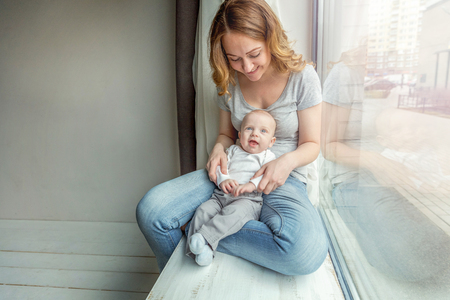 Young mother holding newborn child. Woman and infant boy relax and playing in bedroom near windiow. Mom of breast feeding baby. Family, maternity, tenderness, parenthood, responsibility conceptの写真素材