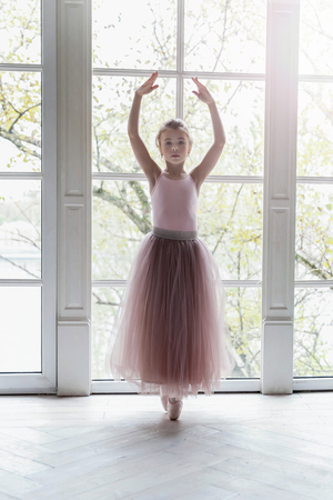 Young classical ballet dancer girl in dance class. Beautiful graceful ballerine practice ballet positions in pink tutu skirt near large window in white light hallの写真素材