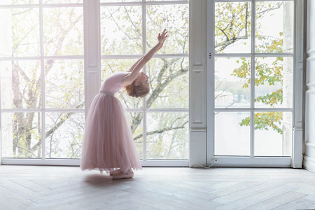 Young classical ballet dancer girl in dance class. Beautiful graceful ballerine practice ballet positions in pink tutu skirt near large window in white light hallの写真素材