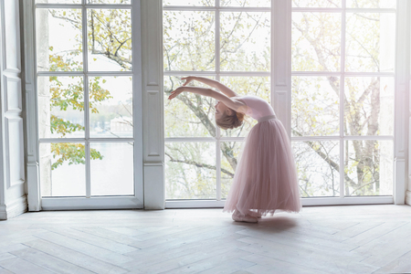 Young classical ballet dancer girl in dance class. Beautiful graceful ballerine practice ballet positions in pink tutu skirt near large window in white light hallの写真素材