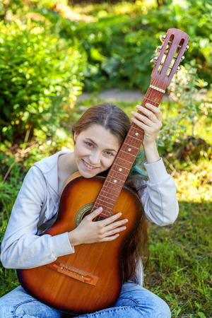Young hipster woman sitting in grass and playing guitar on park or garden background. Teen girl learning to play song and writing music. Hobby, lifestyle, relax, Instrument, leisure, education conceptの写真素材