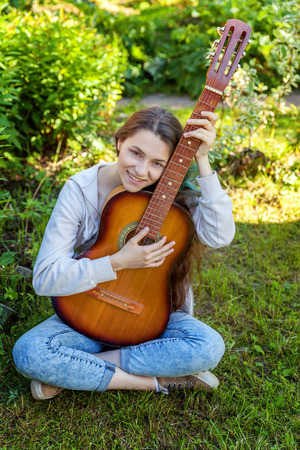 Young hipster woman sitting in grass and playing guitar on park or garden background. Teen girl learning to play song and writing music. Hobby, lifestyle, relax, Instrument, leisure, education conceptの写真素材