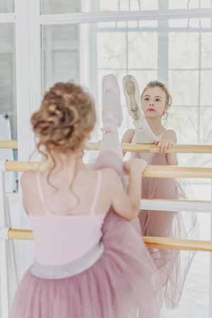 Young classical ballet dancer girl in dance class. Beautiful graceful ballerina practice ballet positions in pink tutu skirt near large mirror in white light hallの写真素材