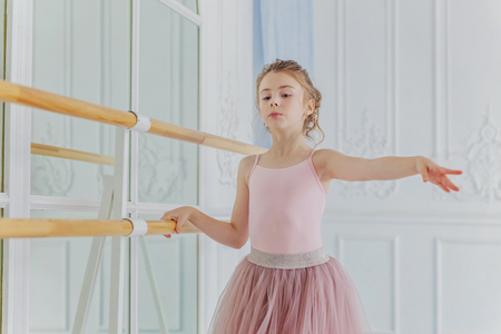 Young classical ballet dancer girl in dance class. Beautiful graceful ballerina practice ballet positions in pink tutu skirt near large mirror in white light hallの写真素材