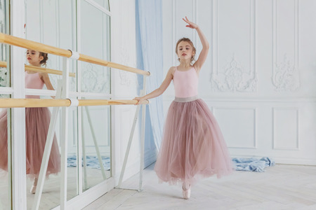 Young classical ballet dancer girl in dance class. Beautiful graceful ballerina practice ballet positions in pink tutu skirt near large mirror in white light hallの写真素材
