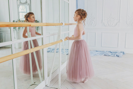 Young classical ballet dancer girl in dance class. Beautiful graceful ballerina practice ballet positions in pink tutu skirt near large mirror in white light hallの写真素材
