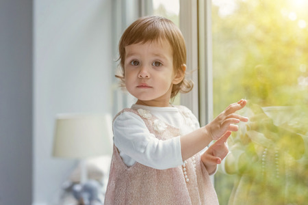 Little cute sweet smiling girl in pastel pink dress standing on the window sill in bright light living room at home and smiling. Childhood, preschool, youth, relax conceptの写真素材