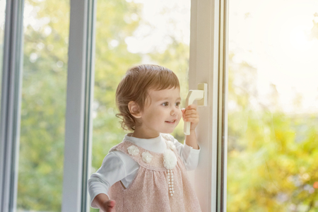 Little cute sweet smiling girl in pastel pink dress standing on the window sill in bright light living room at home and smiling. Childhood, preschool, youth, relax conceptの写真素材