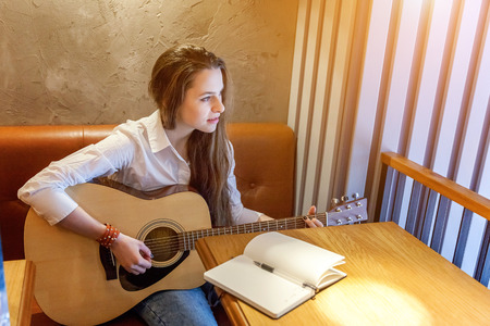 Young hipster woman sitting at table and playing guitar in cafe background. Teen girl learning to play song and writing music. Hobby, lifestyle, relax, Instrument, leisure, education conceptの写真素材