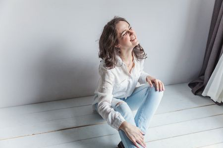 Happy teenage girl smiling. Young happy positive woman in white shirt sitting on floor in bright room against white wall. European woman. Positive human emotion facial expression body languageの写真素材