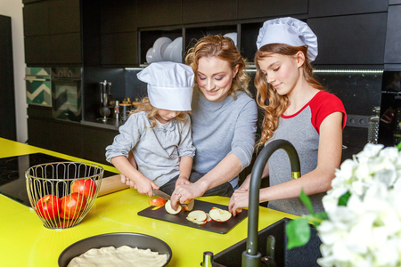 Happy family in kitchen. Mother and two children preparing dough, bake apple pie. Mom and daughters cooking healthy food at home and having fun. Household, teamwork helping, maternity conceptの写真素材