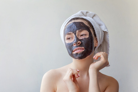 Beauty portrait of a smiling brunette woman in a towel on the head applying black nourishing mask on face on white background isolated. Skincare cleansing spa relax cosmetics conceptの写真素材