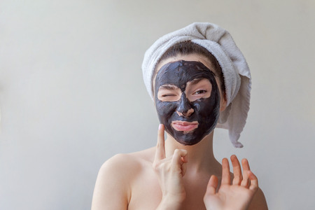 Beauty portrait of a smiling brunette woman in a towel on the head applying black nourishing mask on face on white background isolated. Skincare cleansing spa relax cosmetics conceptの写真素材