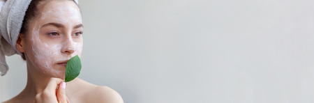 Beauty portrait of woman in towel on head with white nourishing mask or creme on face and green leaf in hand, white background isolated. Skincare cleansing eco organic cosmetic spa concept Bannerの写真素材