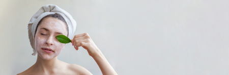 Beauty portrait of woman in towel on head with white nourishing mask or creme on face and green leaf in hand, white background isolated. Skincare cleansing eco organic cosmetic spa concept Bannerの写真素材