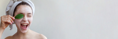 Beauty portrait of woman in towel on head with white nourishing mask or creme on face and green leaf in hand, white background isolated. Skincare cleansing eco organic cosmetic spa concept Bannerの写真素材