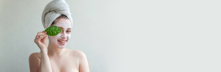 Beauty portrait of woman in towel on head with white nourishing mask or creme on face and green leaf in hand, white background isolated. Skincare cleansing eco organic cosmetic spa concept Bannerの写真素材