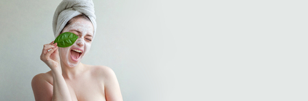 Beauty portrait of woman in towel on head with white nourishing mask or creme on face and green leaf in hand, white background isolated. Skincare cleansing eco organic cosmetic spa concept Bannerの写真素材