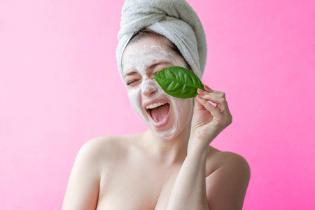 Beauty portrait of a smiling brunette woman in a towel on the head with white nourishing mask or creme on face and green leaf in hand on pink background isolated. Skincare cleansing eco organic cosmetic spa relax conceptの写真素材